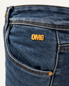 Detalle FV26 Jean Omg: placa OMG dorada, bolsillo delantero, textura denim y costuras.
